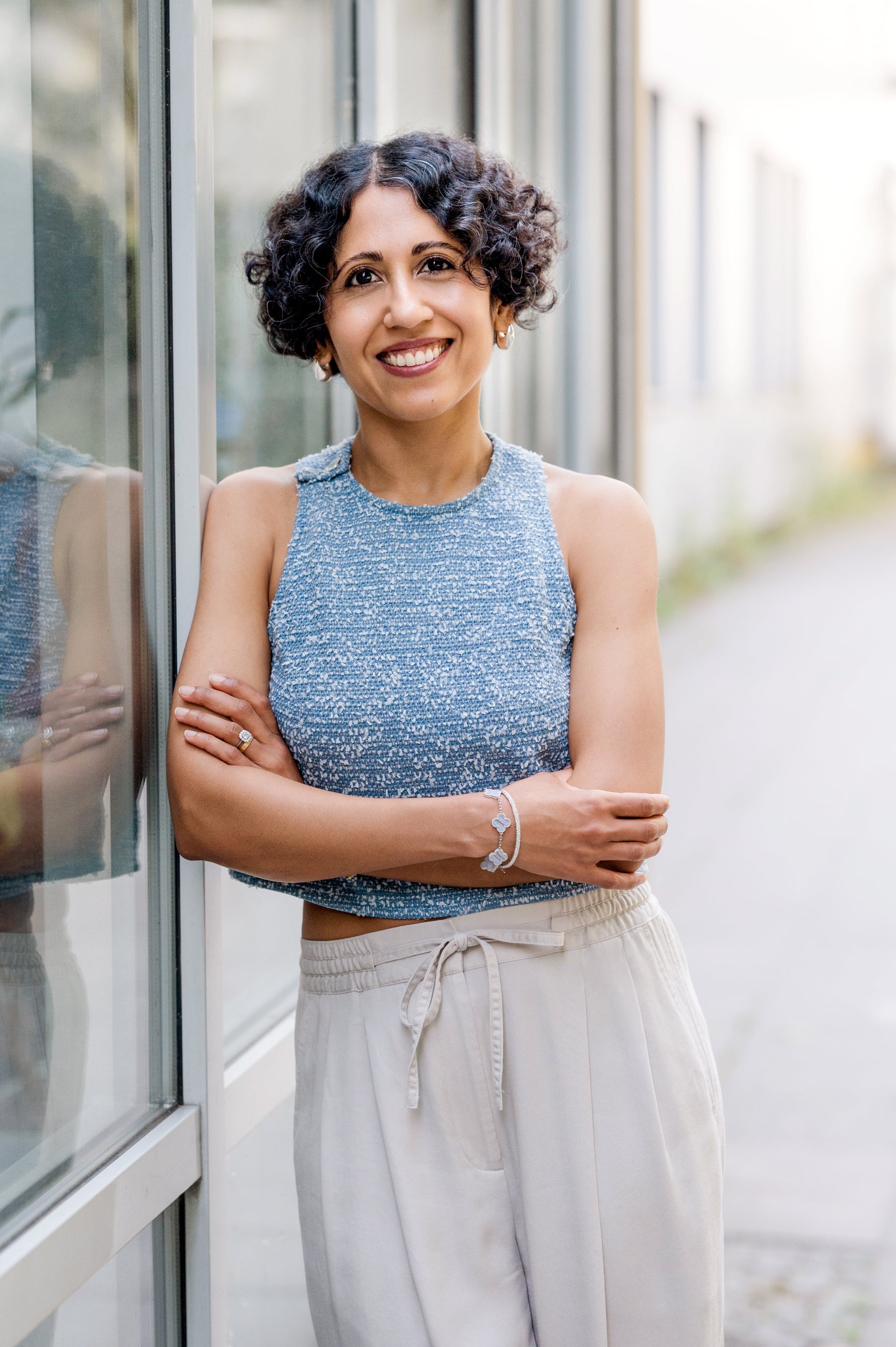 A smiling woman with curly dark hair stands outdoors beside a glass storefront, wearing a sleeveless textured blue top and light beige drawstring pants.